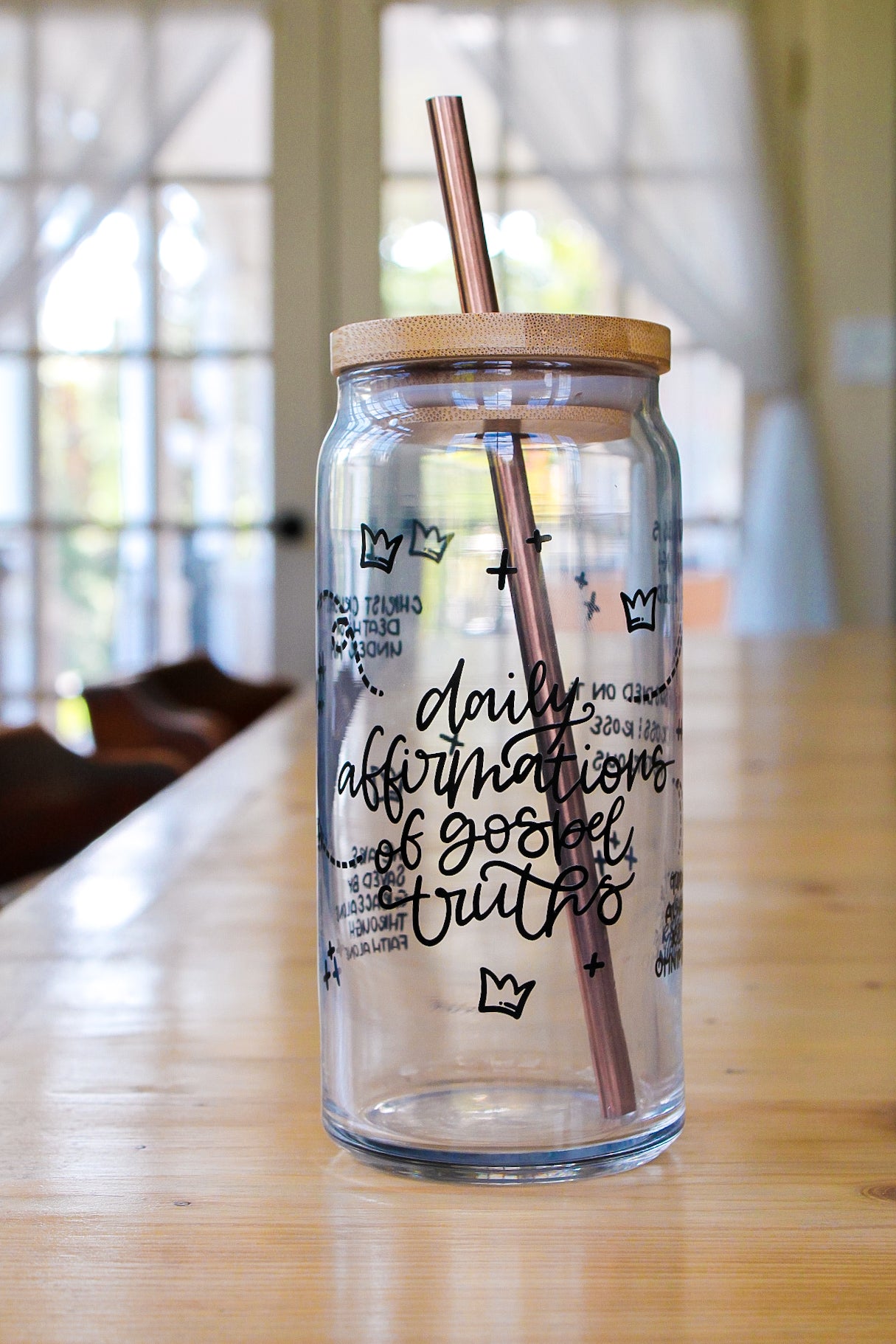 Clear glass tumbler with wooden lid and straw on a wooden surface, featuring gospel truths and crown and cross graphics.