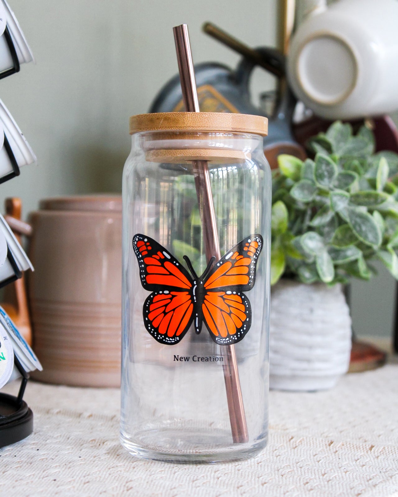 Clear 20 ounce glass can with a butterfly design and wooden lid and metal straw on a coffee bar.