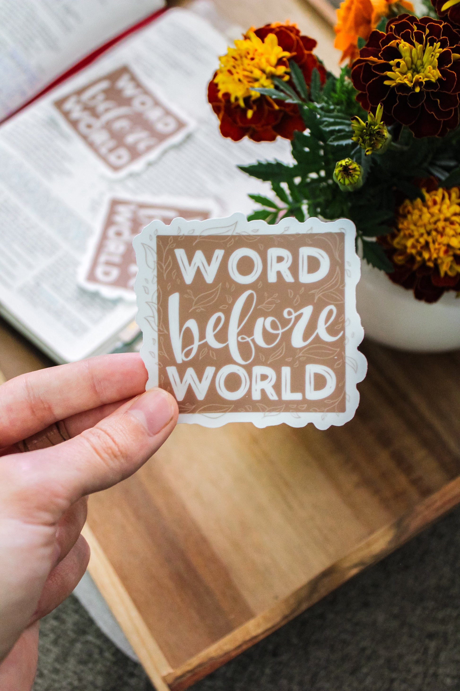 Hand holding a decorative vinyl sticker with 'Word before World' text, flowers in the background