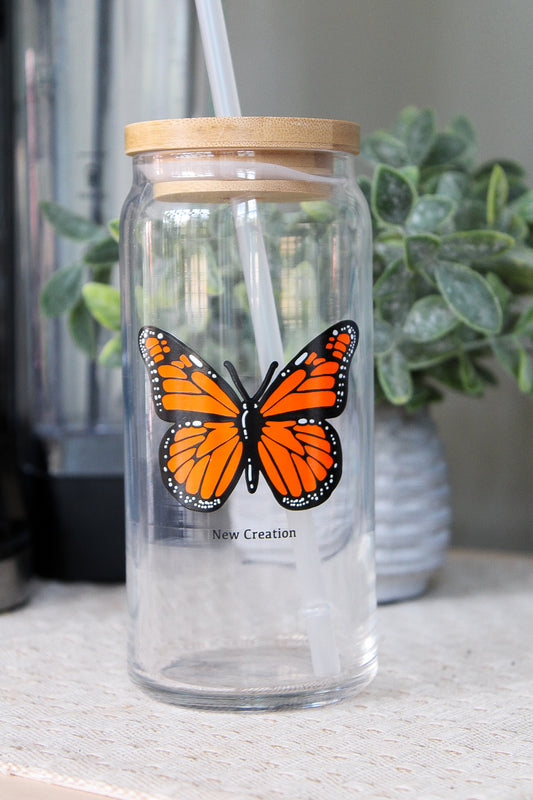 Clear glass tumbler for iced drinks with a butterfly design and wooden lid on a surface with plants in the background.