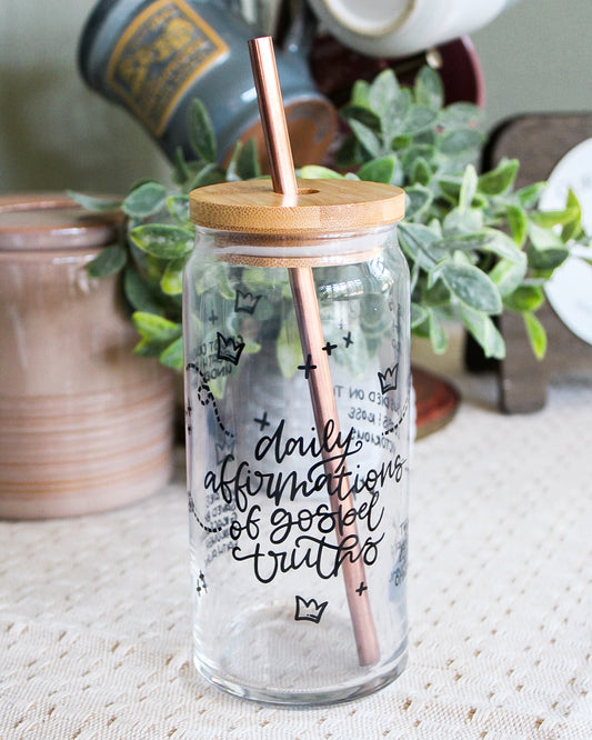 Glass jar with bamboo lid and rose gold straw, containing 'daily affirmations of gospel truths' text, on a coffee bar with plants in the background.