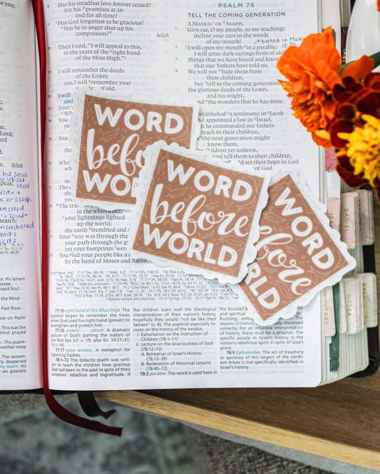 Open Bible with 'Word before World' hand-lettered stickers and flowers on a wooden surface